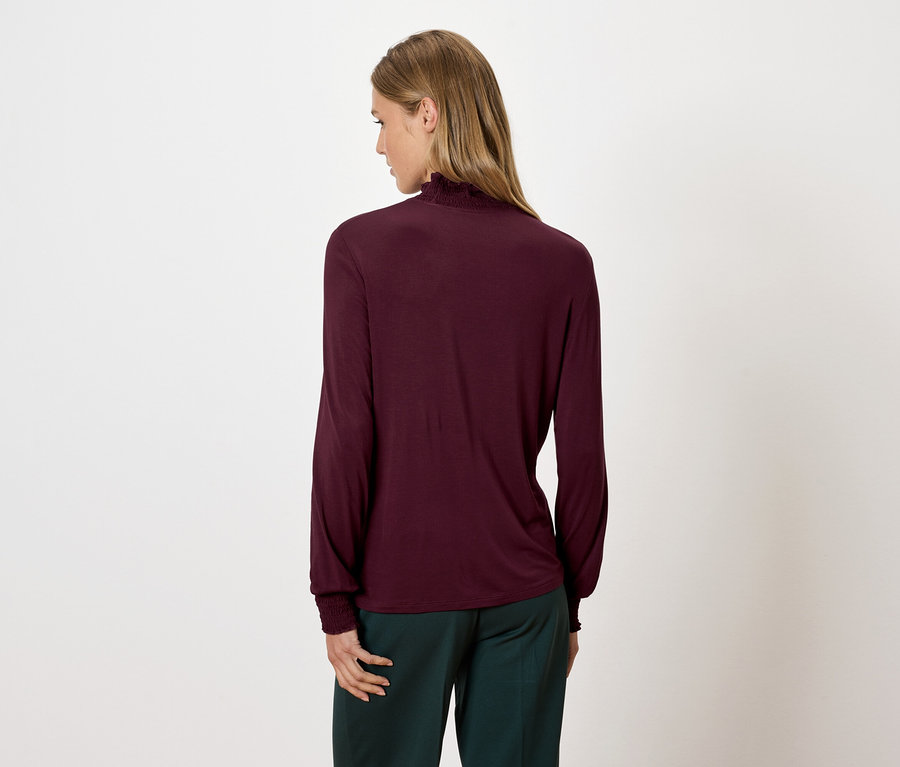 Nő visel bordó, hosszú ujjú garbót és zöld nadrágot, hátulnézetből.