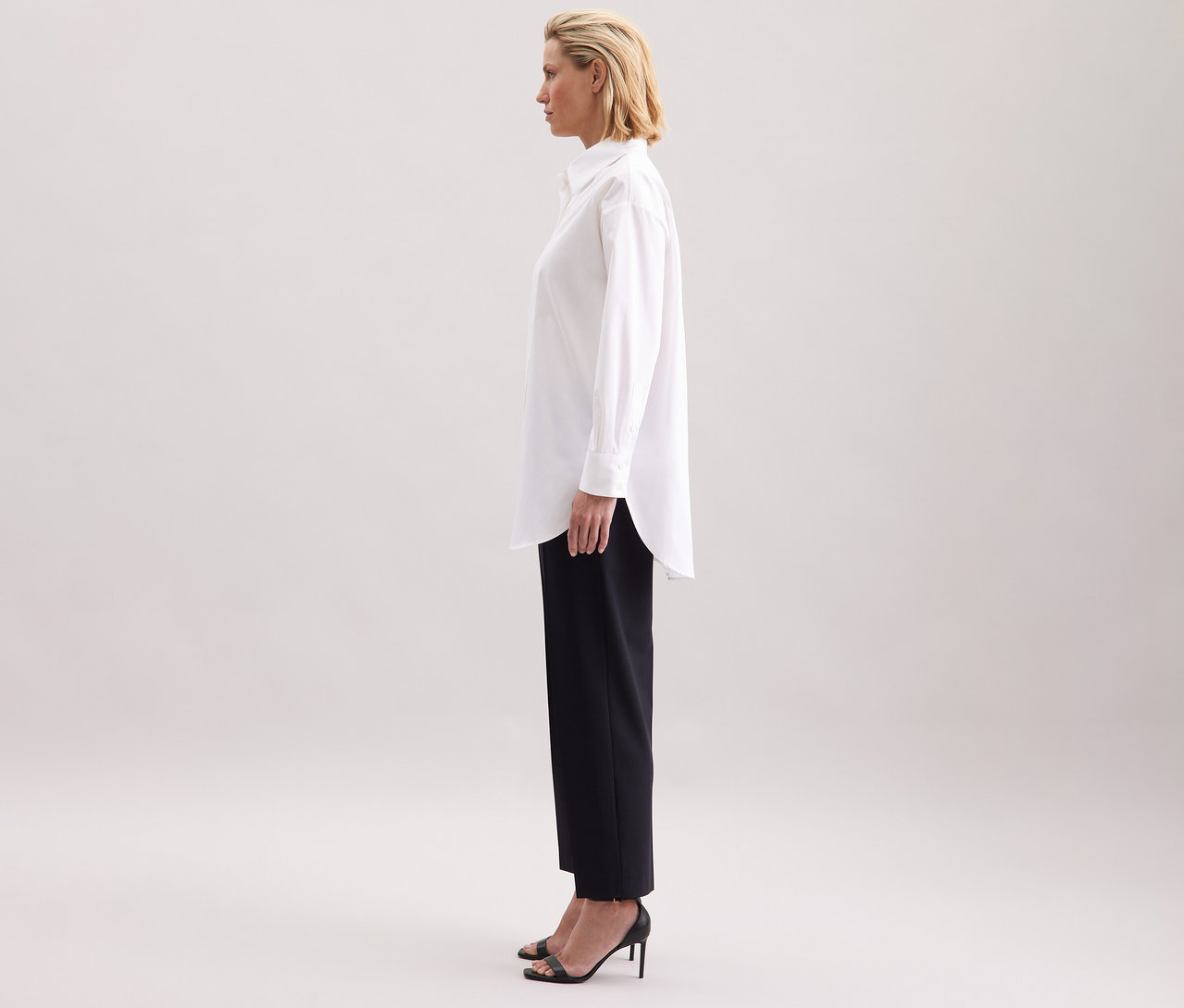 Nő visel egy fehér Seidensticker túlméretes hosszú ingblúzt, fekete nadrágot és magas sarkú cipőt.