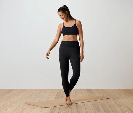 Nő áll egy jógaszőnyegen, fekete sportmelltartót és fekete leggingset visel.