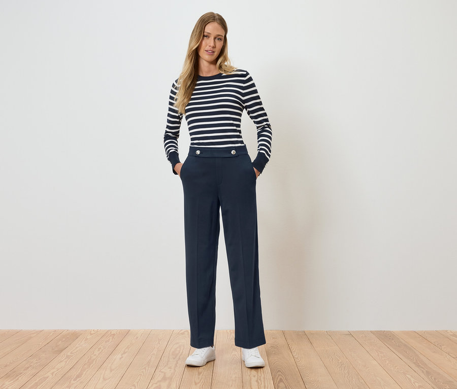 Egy nő áll, kezei a zsebében. Csíkos, hosszú ujjú felsőt és sötétkék nadrágot visel.