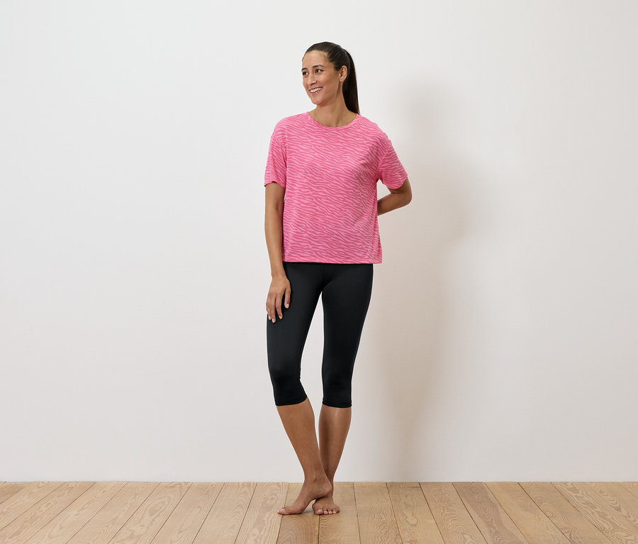 Egy nő áll, rózsaszín pólót és fekete leggingset visel.