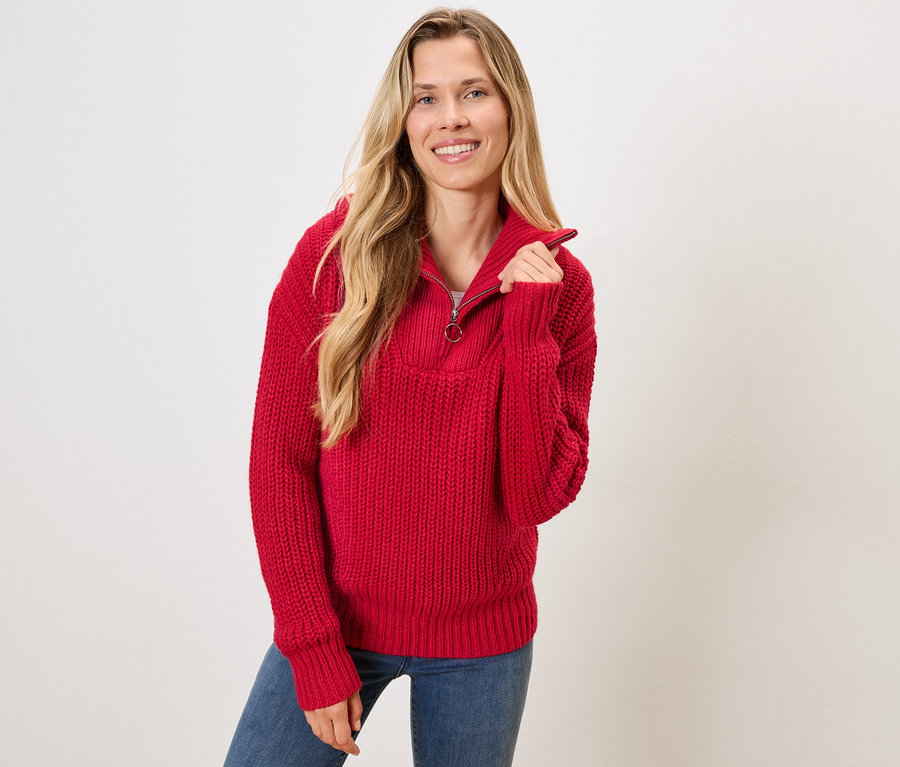 Egy szőke hajú nő piros pulóvert és kék farmert visel.