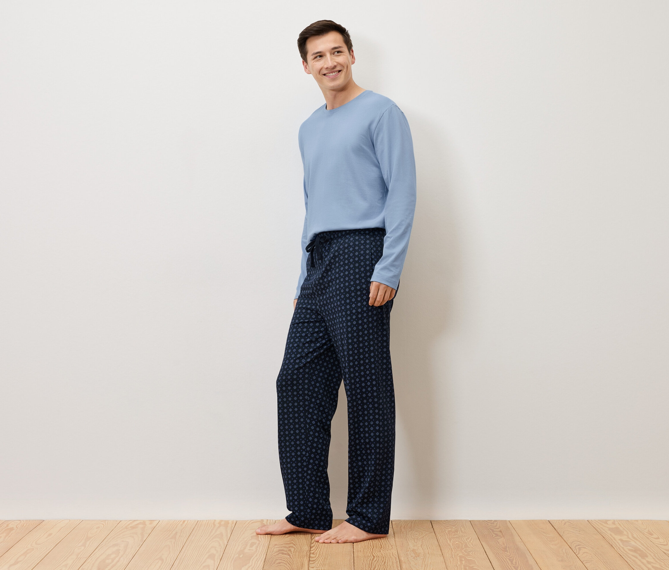 Egy férfi mezítláb áll egy fehér fal előtt, világoskék pulóvert és sötétkék, pöttyös pizsamanadrágot visel.