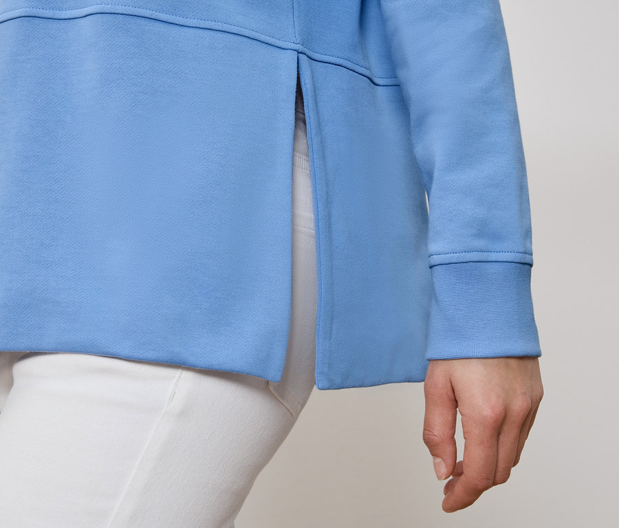 Egy személy világoskék színű, oldalt sliccelt pulóvert és fehér nadrágot visel.