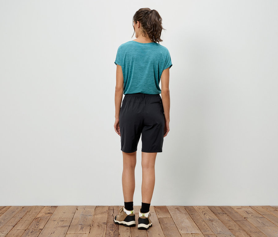 Hátulnézet egy lófarokos nőről, aki türkizkék pólót, fekete rövidnadrágot és tornacipőt visel egy fapadlón.