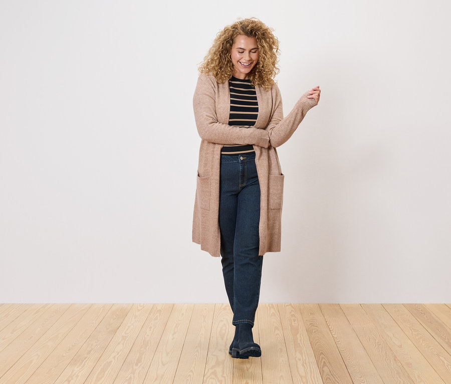 A göndör hajú nő hosszú, világosbarna kardigánt visel egy fekete-fehér csíkos felső és sötét farmer felett.