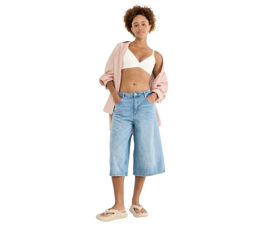 Nő áll zsebre tett kézzel, fehér bralette-et, rózsaszín csíkos blúzt, világoskék culotte-ot és bézs papucsot visel.