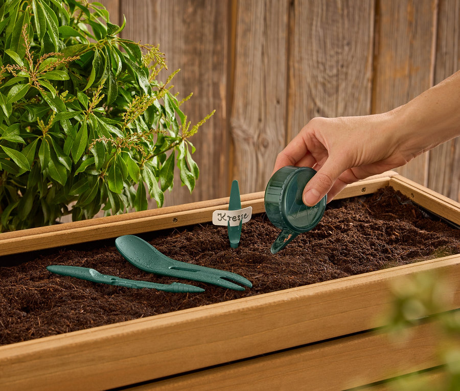 Egy kéz vizet önt egy kis zöld locsolókannából egy földdel teli fadobozba, amelyben zöld kerti szerszámok és egy "Kresse" feliratú címke található.