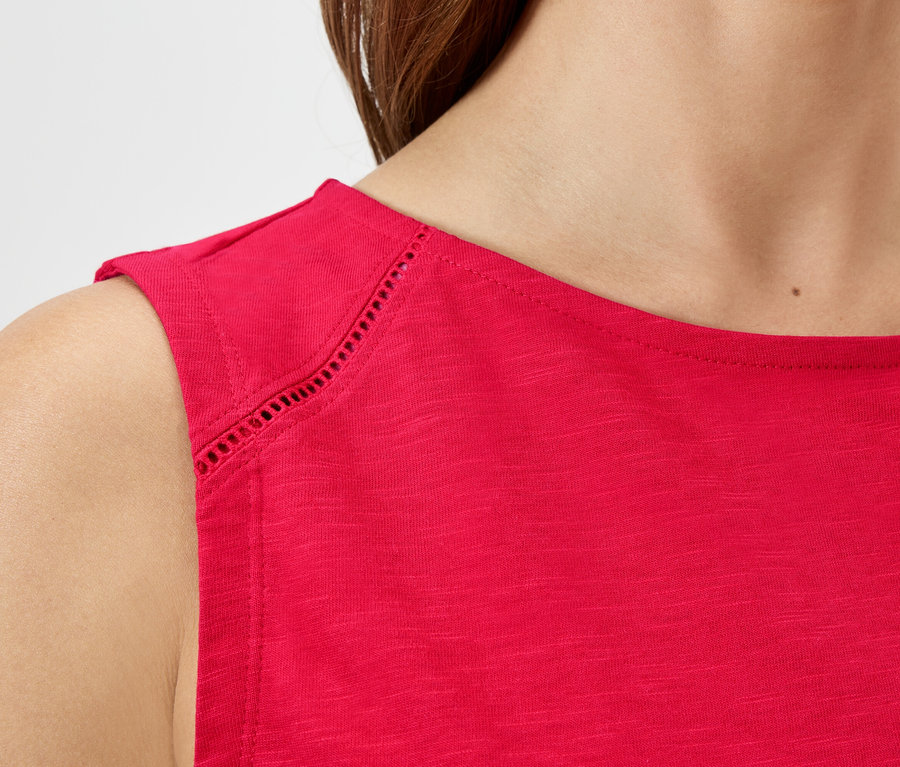 Közeli felvétel egy piros, ujjatlan felsőt viselő nőről, melynek vállán lyukacsos minta látható.