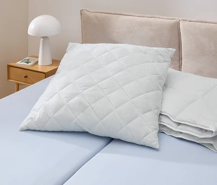 Egy világoskék ágyon egy fehér, steppelt Cell Solution® párna (90 x 70 cm) fekszik. Mellette más Cell Solution® párnák (80 x 80 cm, 100 x 65 cm) találhatók.
