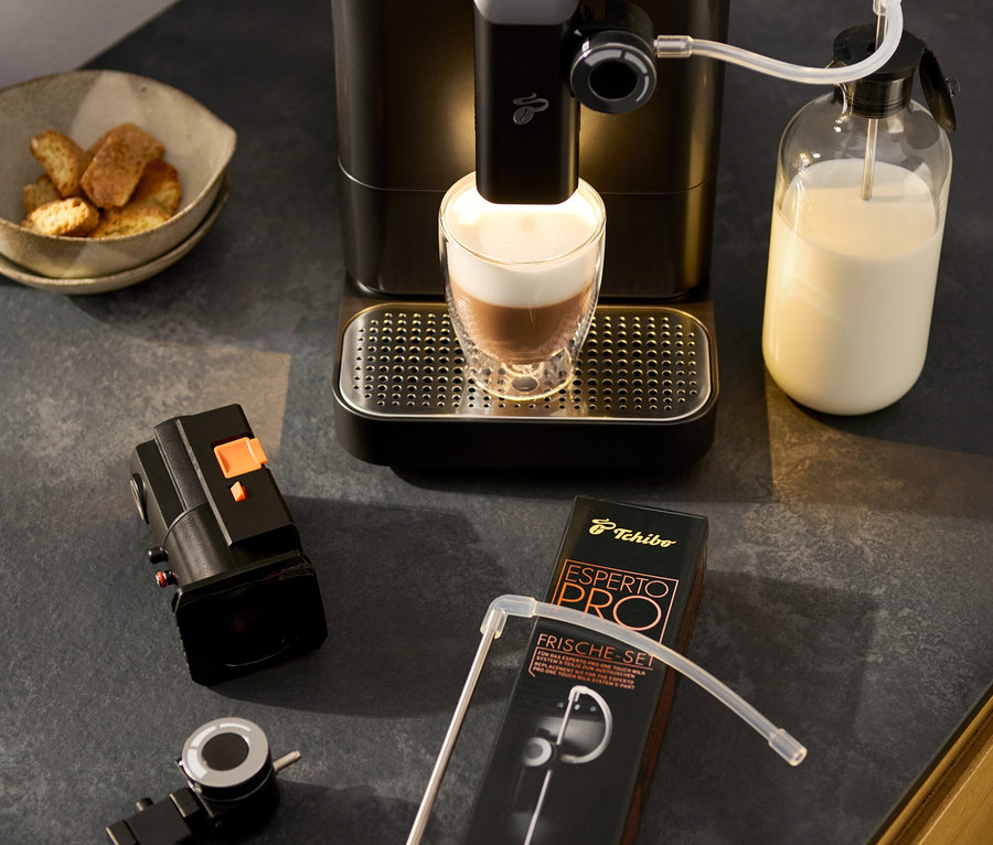 A »Esperto 2 Milk« Granite Black kávéfőző latte macchiatót készít. Mellette egy tejhab forgatószabályzó, egy tál sütemény és egy tartozékkészlet csomagolása található.