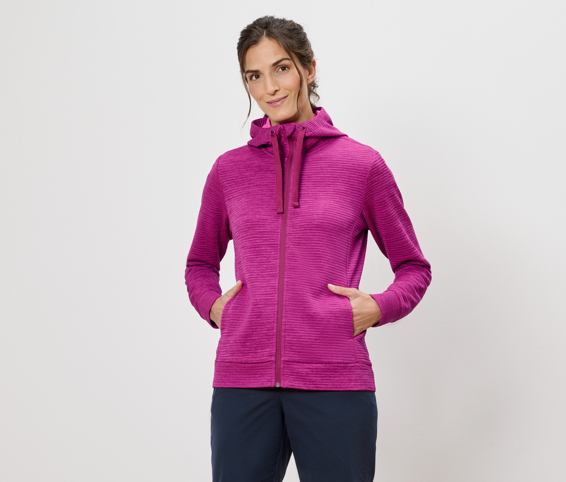 Nő rózsaszín kapucnis pulóverben, kezével a zsebében, fehér háttér előtt.