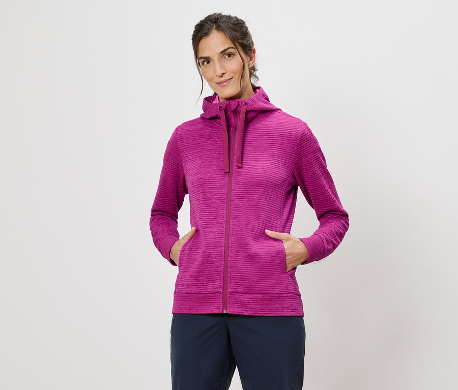 Nő rózsaszín kapucnis pulóverben, kezével a zsebében, fehér háttér előtt.