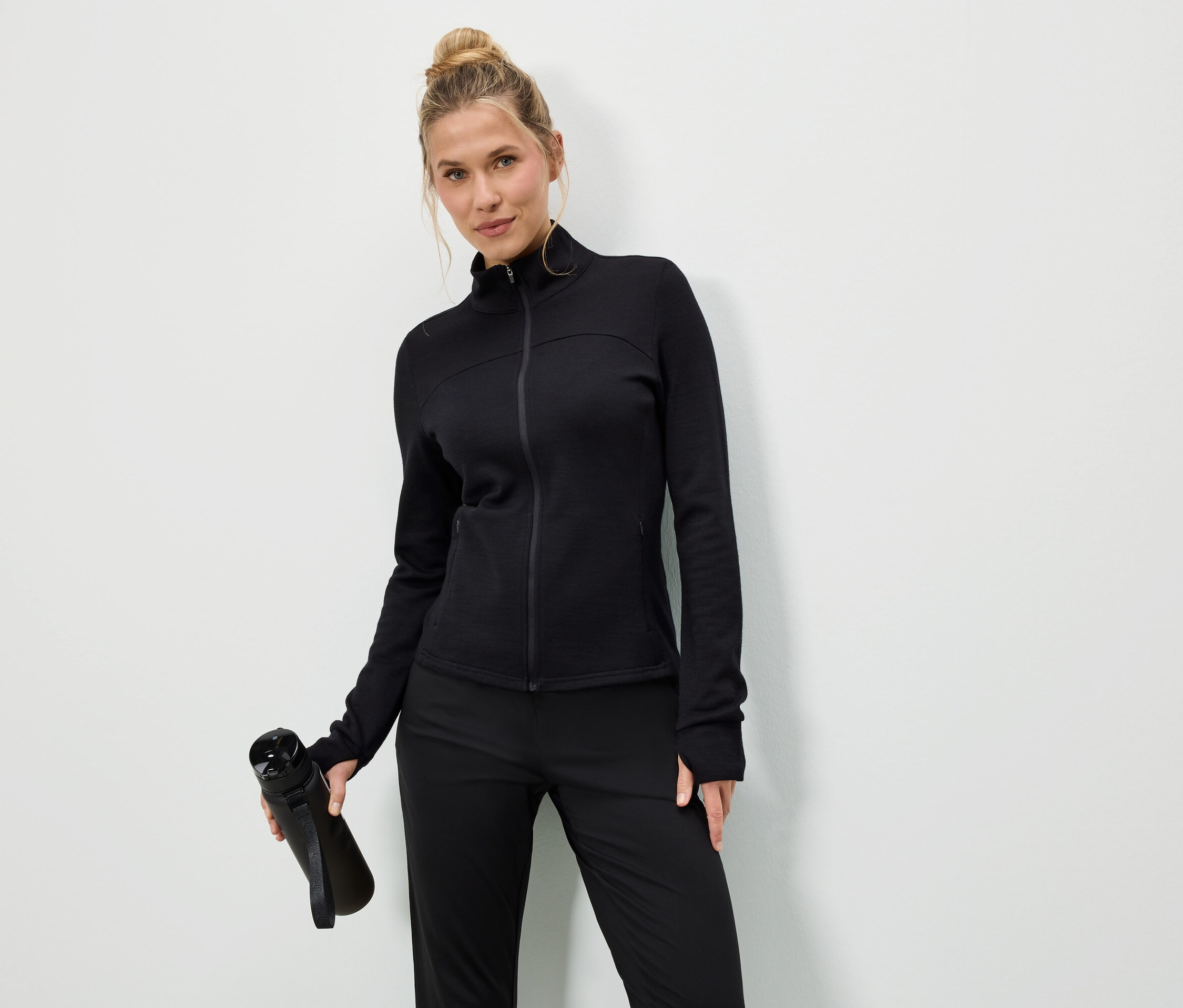 Nő fekete kabátban és nadrágban fekete vizes palackot tart fehér háttér előtt.