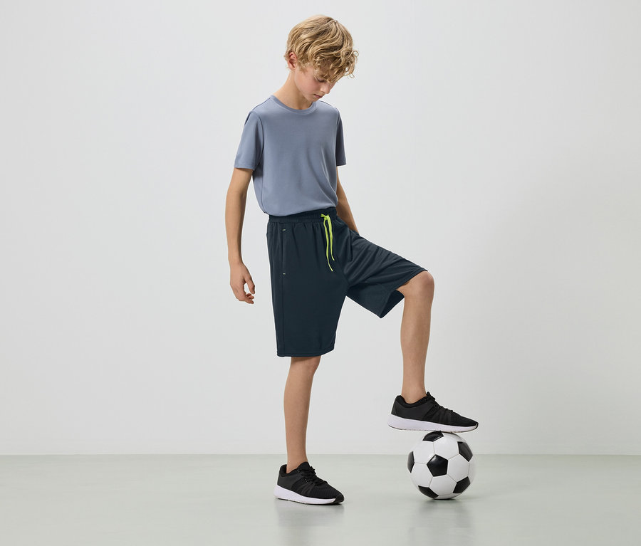 Egy szőke fiú szürke pólóban és sötét rövidnadrágban, egyik lábával egy focilabdán áll.