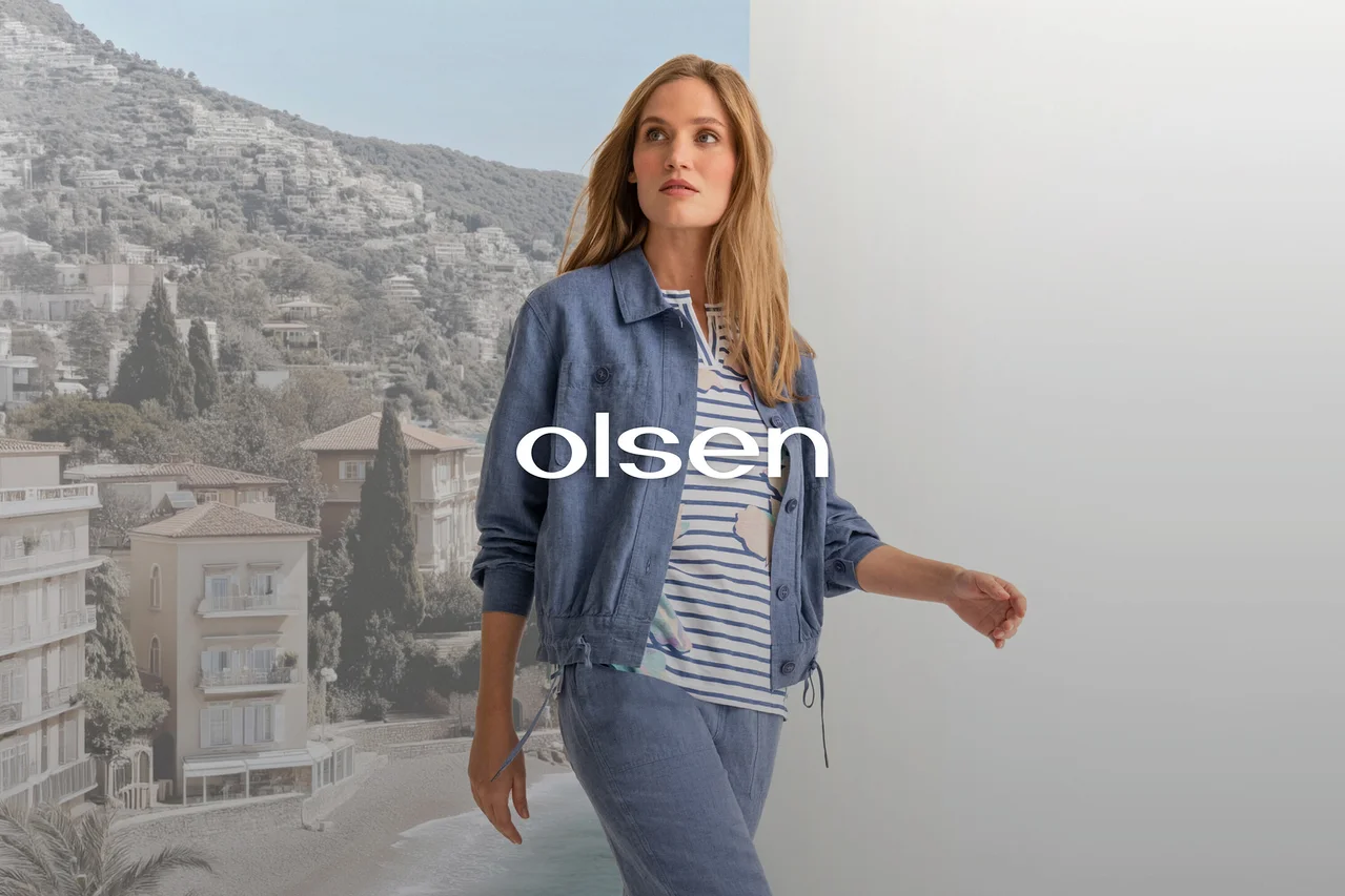 Nő kék dzsekiben, csíkos felsőben és kék nadrágban, egy tengerparti város és egy szürke fal osztott háttere előtt sétál, 'olsen' felirattal.