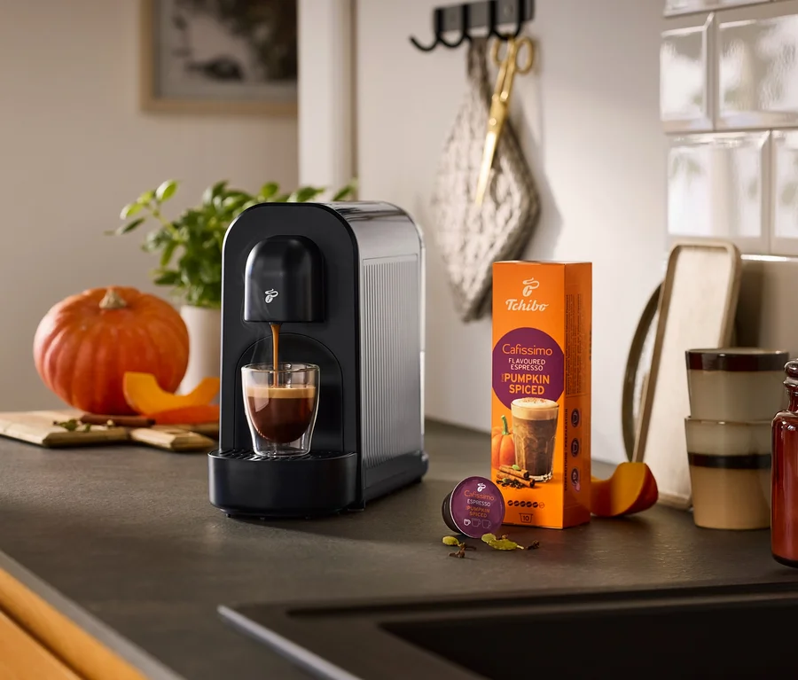 Fekete Cafissimo now kávéfőző készít espressót egy pohárba, mellette egy Cafissimo Pumpkin Spiced kávé csomag és egy kapszula, tökkel és fűszerekkel körülvéve egy konyhapulton.