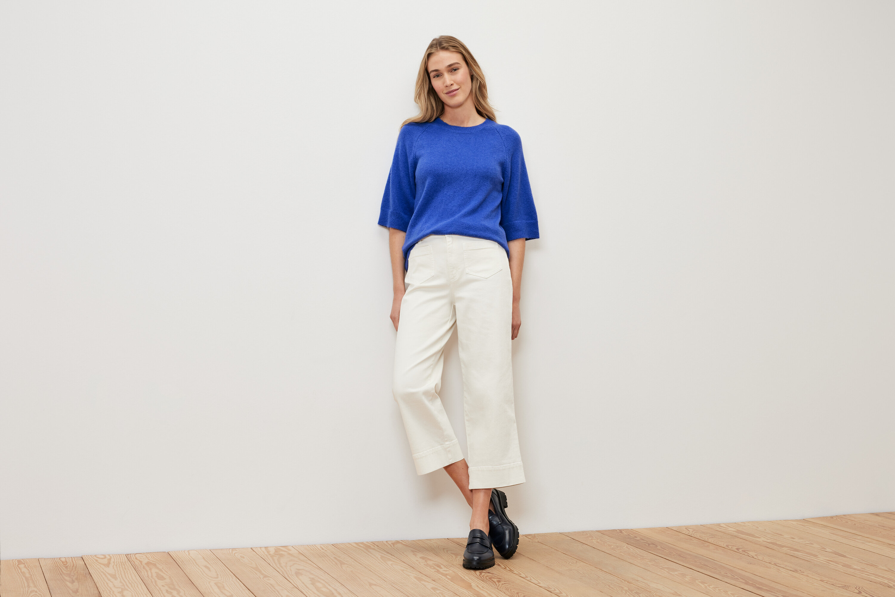 Egy nő áll egy fehér fal előtt, kobaltkék kötött pulóvert, fehér nadrágot és fekete cipőt visel.