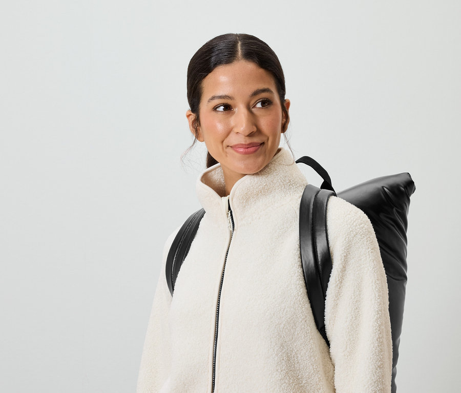 Mosolygó, barna hajú és barna szemű nő krémszínű kabátot és fekete hátizsákot visel.