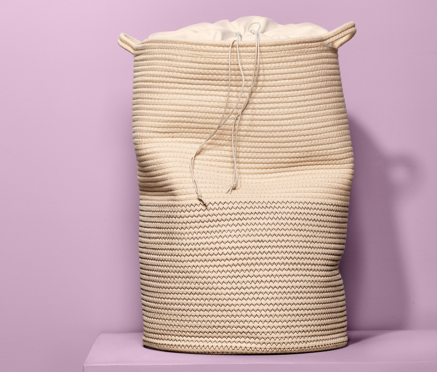 Zsinórral összehúzott szövetbetétes fonott szennyestartó kosár lila háttér előtt, lila talapzaton.
