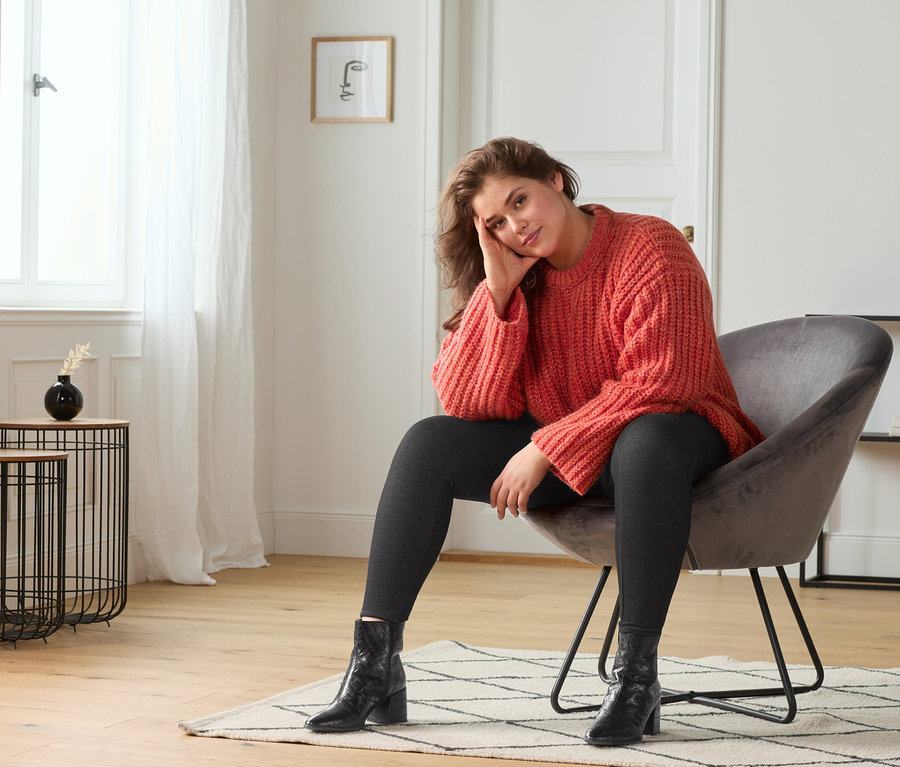 Nő ül egy fotelben, narancspiros kötött pulóvert, farmerhatású leggingset és fekete bőr bokacsizmát visel.