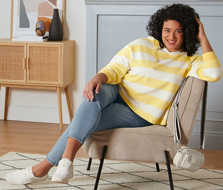 Nő ül egy fotelben, és sárga, csíkos kapucnis szabadidőfelsőt, treggingst, krémszínű velúrbőr sneaker cipőt és krémszínű oldaltáskát visel.