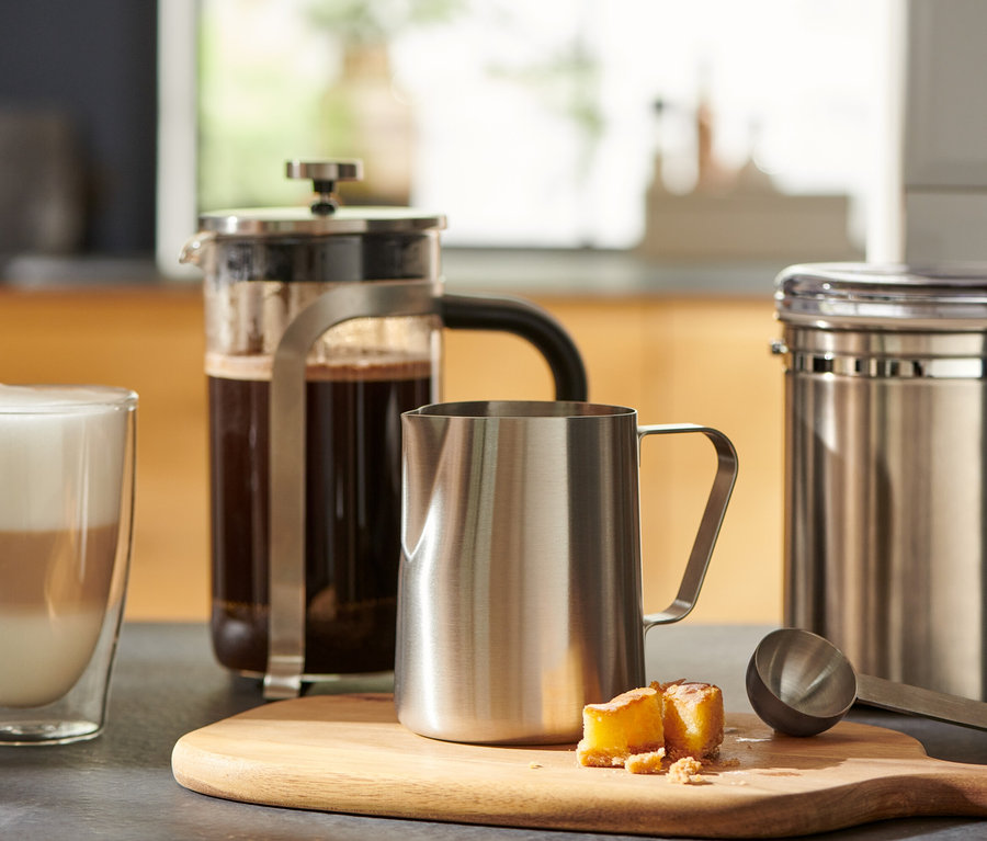 Egy pohár kávé egy french press mellett áll, és egy kis, nemesacélból készült tejeskancsó egy fa vágódeszkán.
