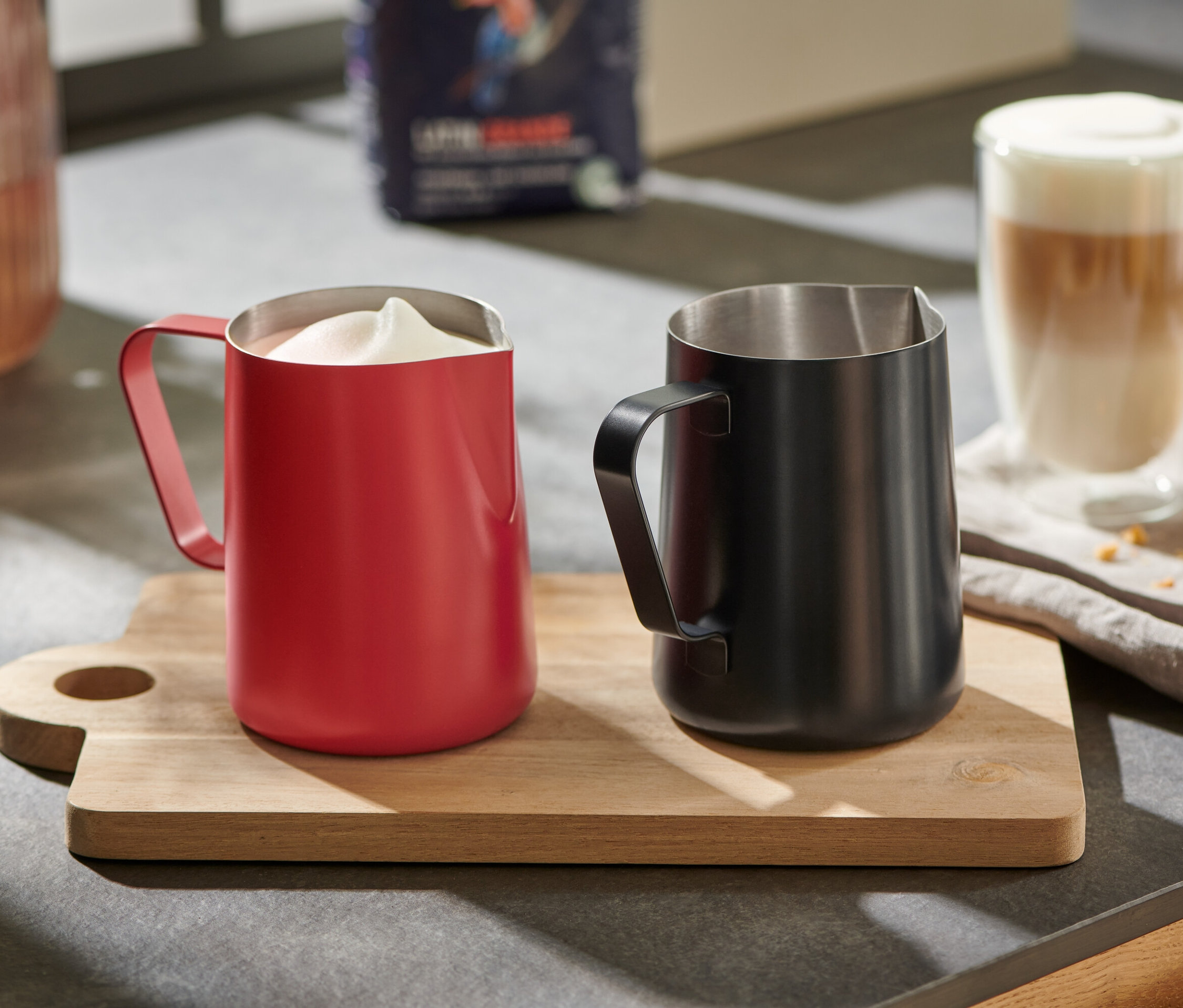 Egy piros tejeskancsó habosított tejjel és egy fekete tejeskancsó egy fa deszkán.