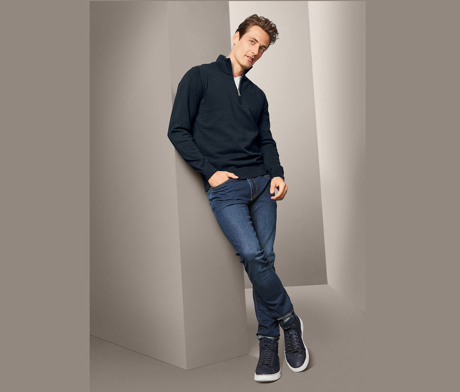 Férfi támaszkodik a falnak, sötétkék férfi állónyakú kötött pulóvert, farmert és tornacipőt visel.