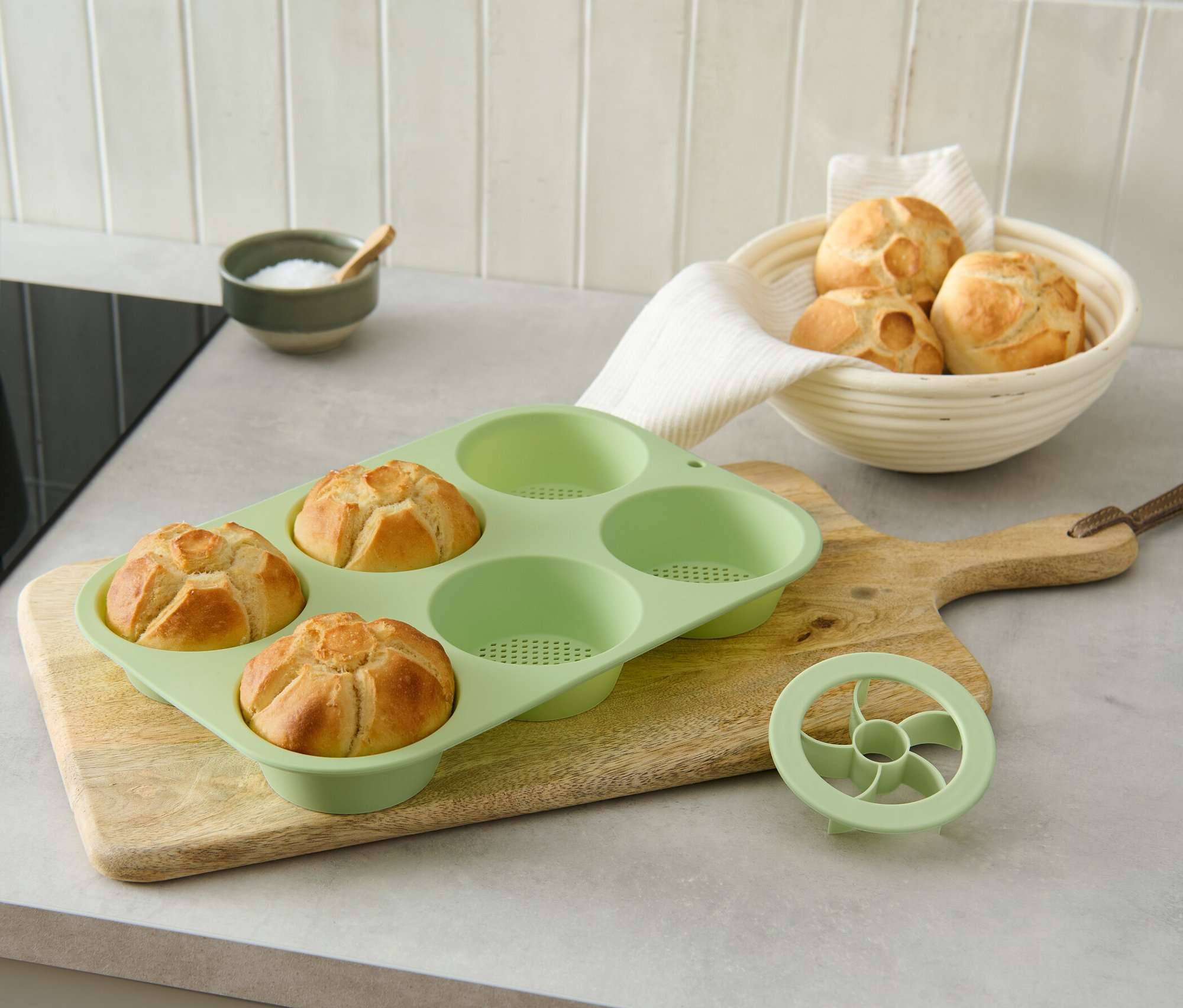 Három sült zsemle egy zöld, perforált sütőforma zsemlékhez egy fából készült vágódeszkán.