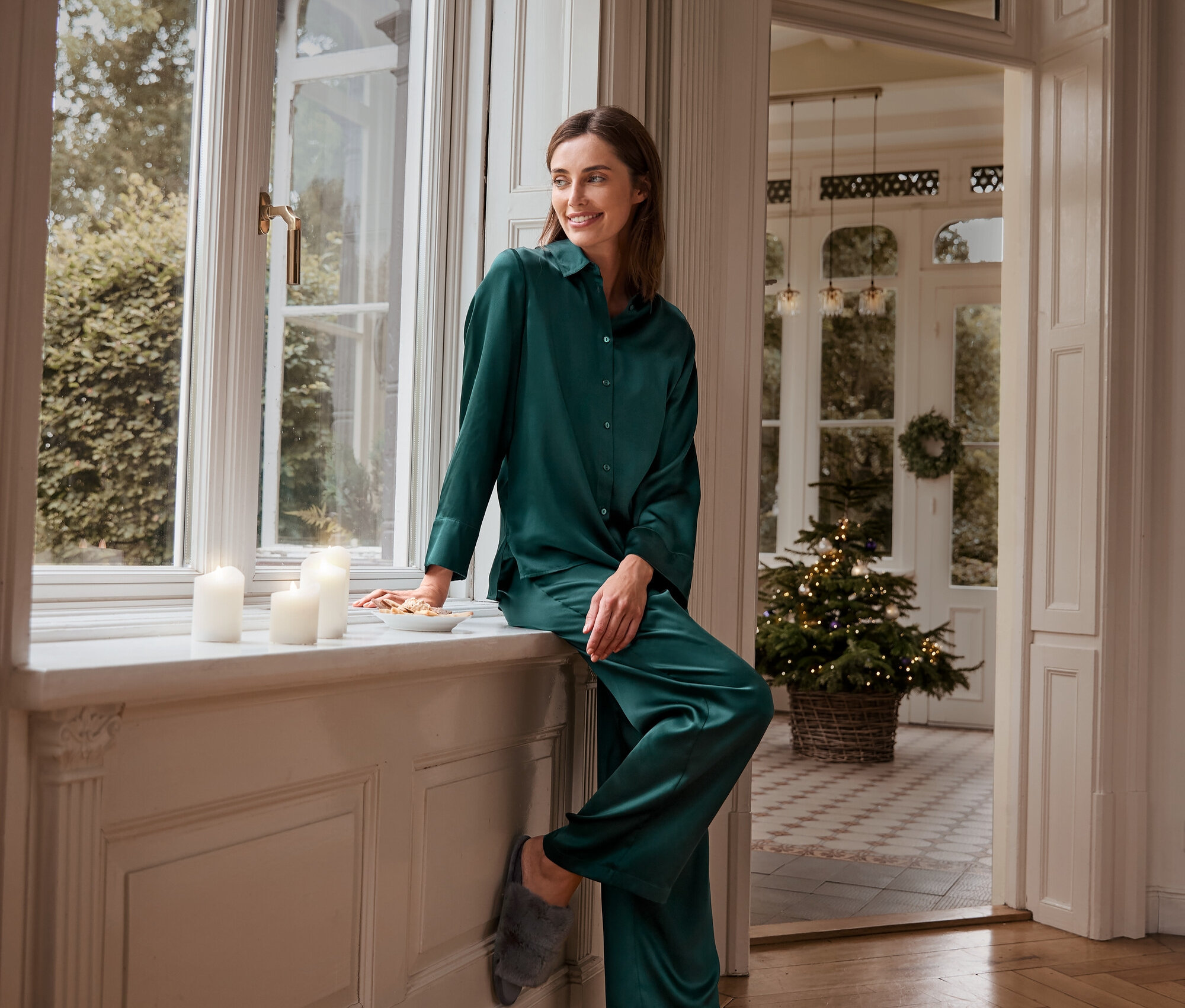 Nő ül az ablakpárkányon sötétzöld selyem pizsamanadrágban, pizsamafelsőben és szürke plüss házipapucsban.