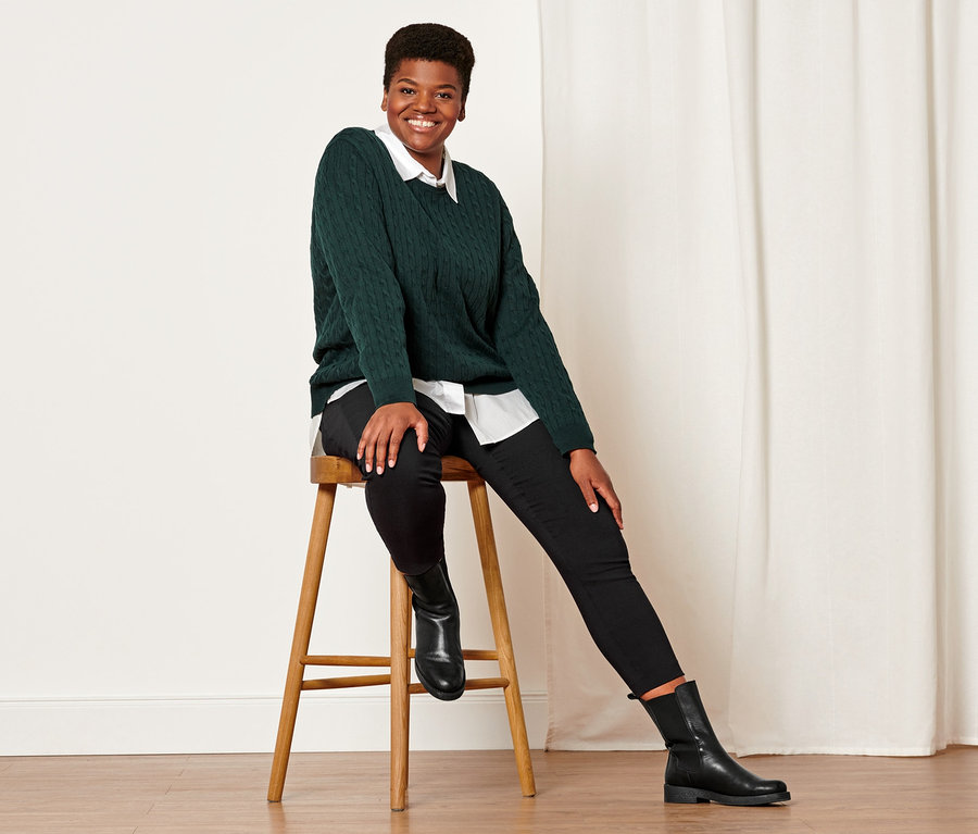 Nő ül egy széken, sötétzöld kötött pulóvert, fekete sztreccsnadrágot és fekete bőrcsizmát visel.