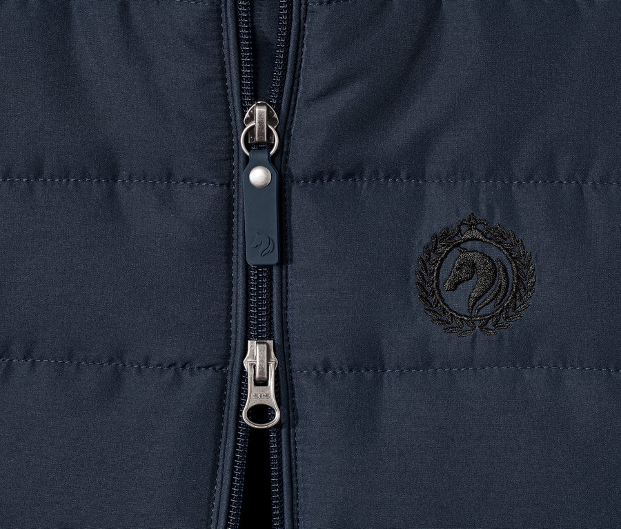 Közeli kép egy sötétkék cipzáras, lovas logóval ellátott lány lovaglómellényről.