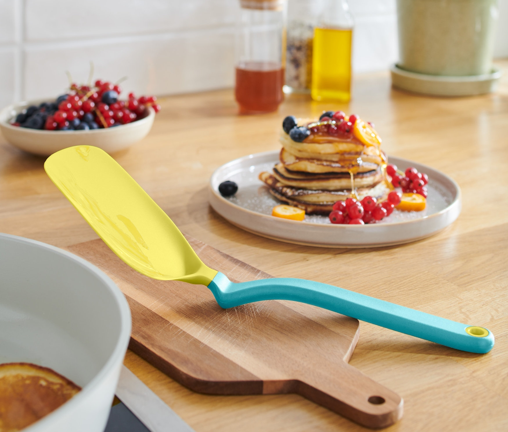 Egy sárga tojás- és palacsintaforgató lapát fekszik egy fa deszkán egy palacsintás serpenyő mellett.