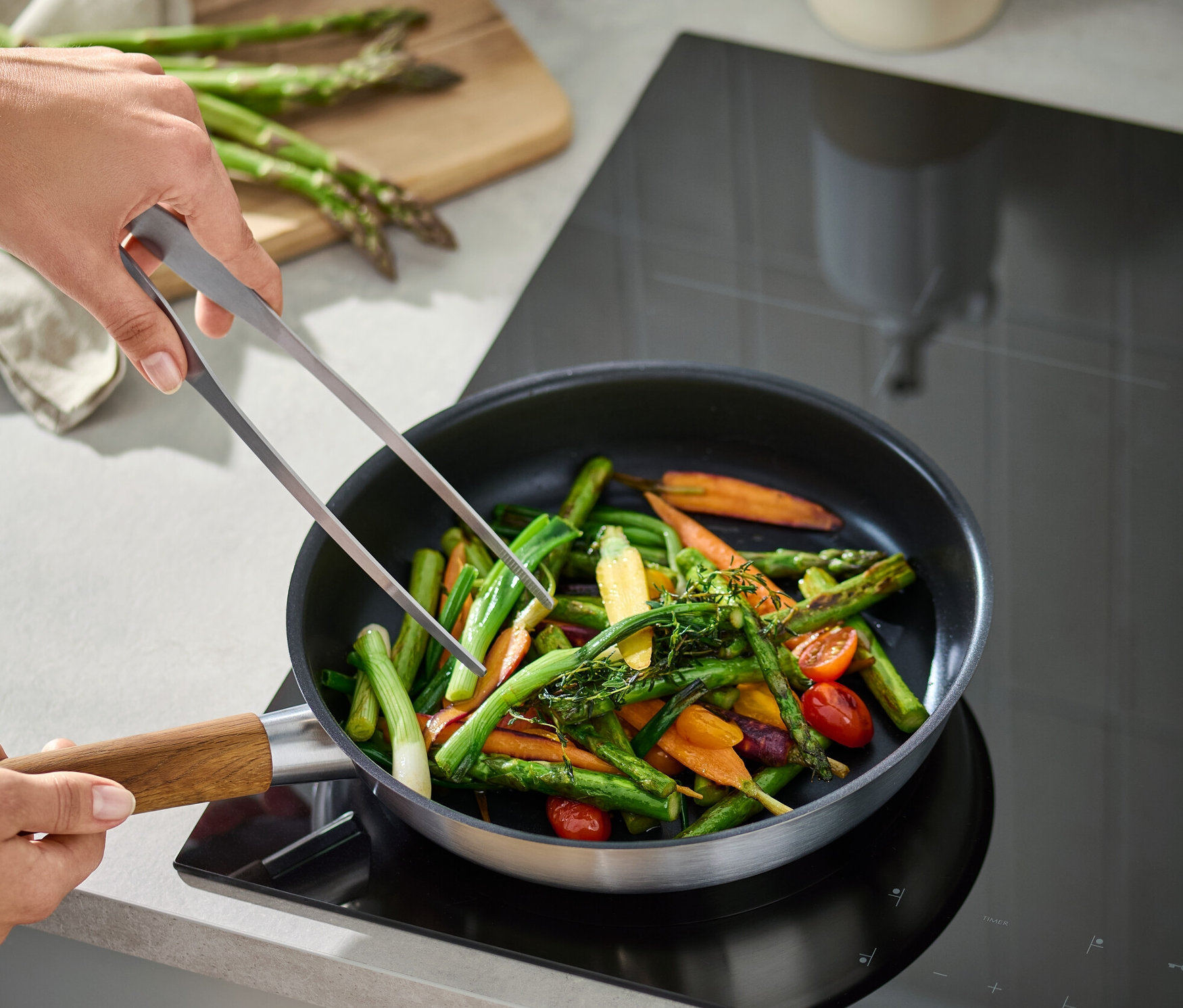 Egy személy nemesacél konyhai csipesszel zöldségeket kever egy serpenyőben.