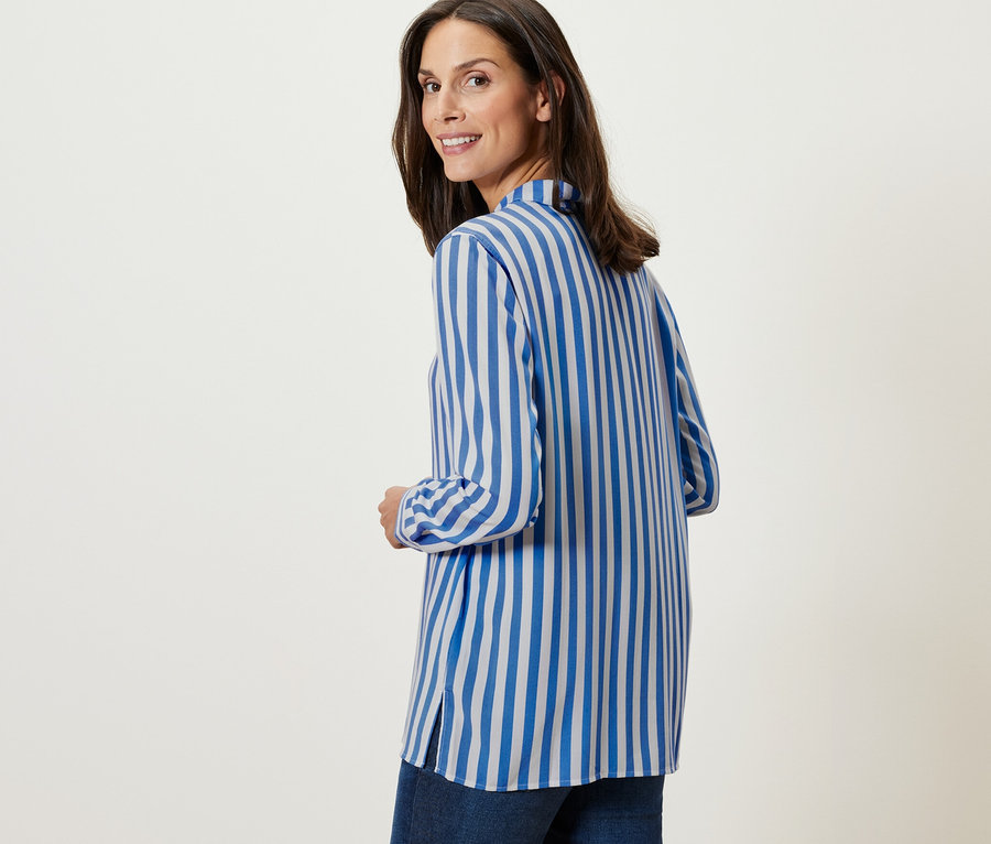 A nő a válla fölött hátra néz, és egy Női ingblúz, csíkos, világoskék/krémfehér színű blúzt visel.