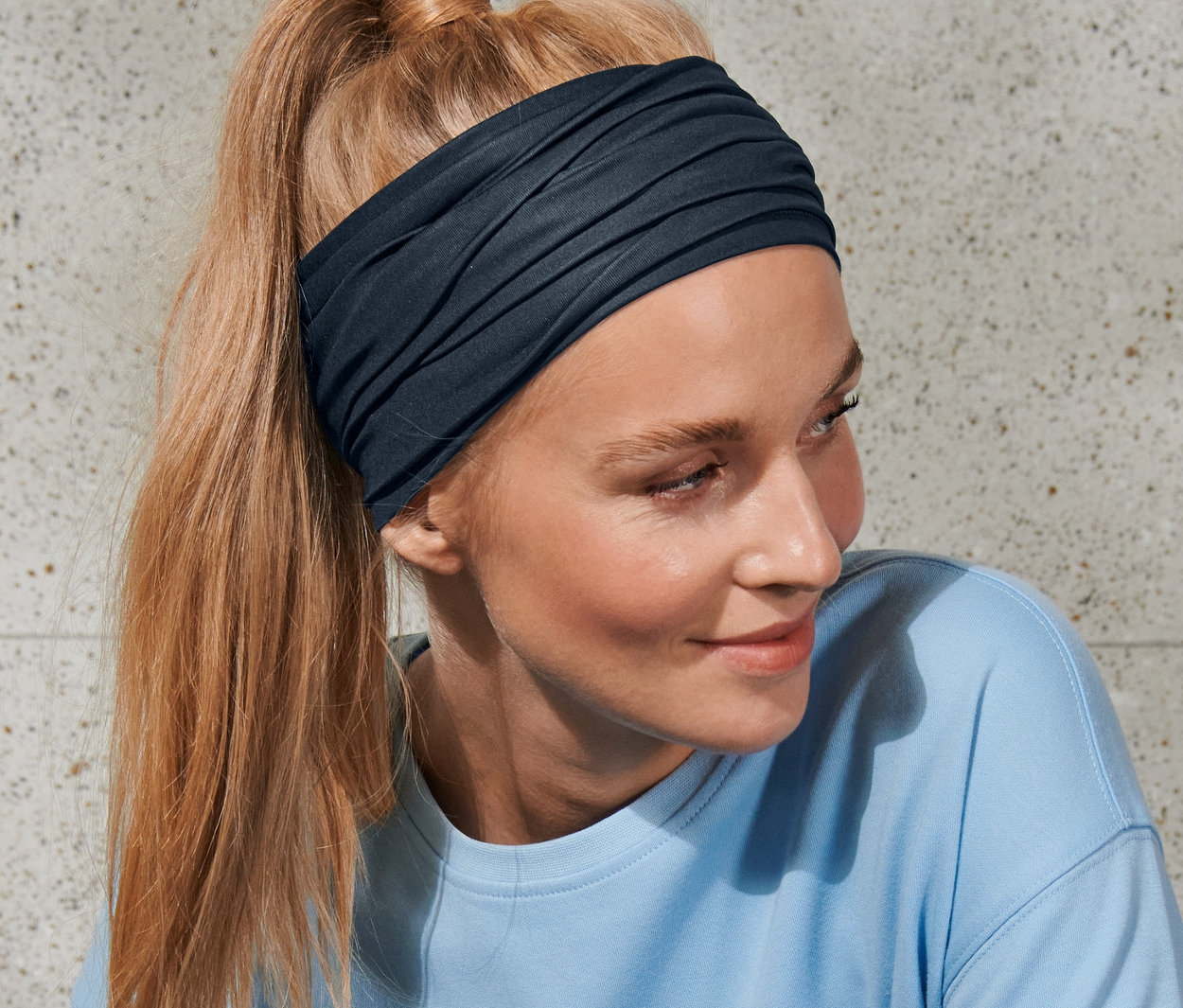 A szőke, lófarokba kötött hajú nő sötétkék/kék unisex multifunkcionális kendőt és kék felsőt visel.