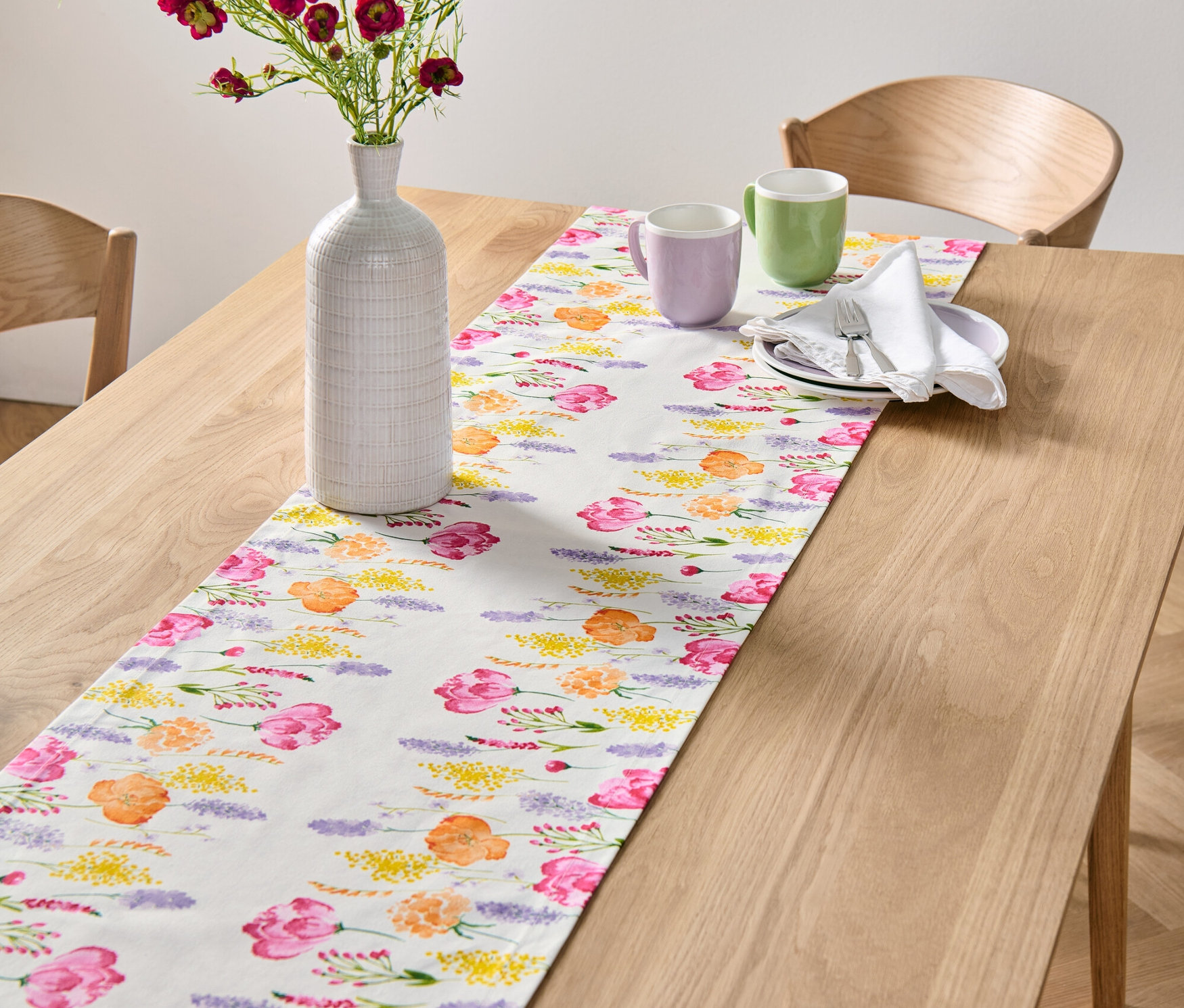 A fa asztalon egy fehér, virágmintás asztali futó található. A futón egy váza virágokkal és két bögre áll.