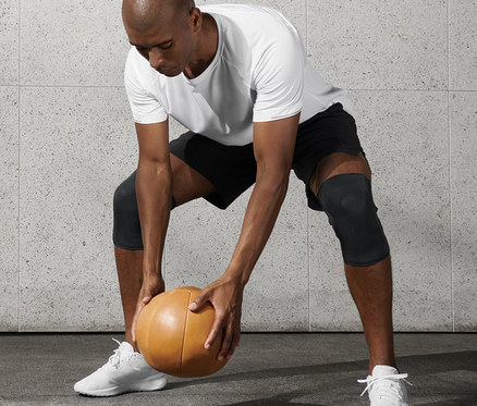 Férfi fehér pólóban és fekete rövidnadrágban medicinlabdát tart, és 2 fitnesz térdvédőt visel.