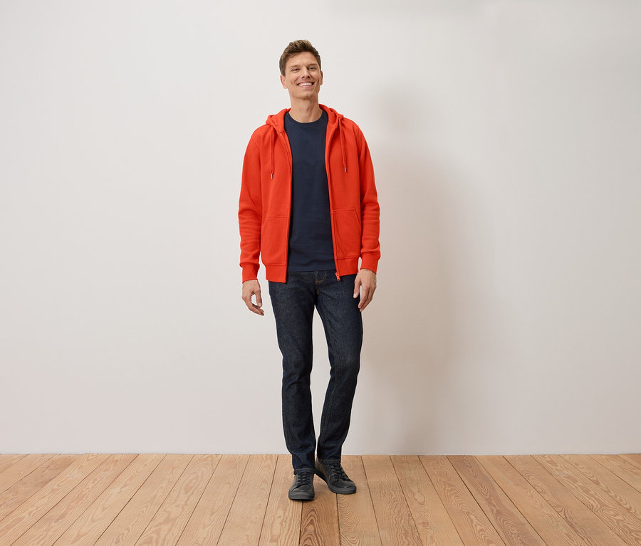 Egy mosolygó férfi áll narancssárga kapucnis pulóverben, sötétkék pólóban és farmerban.