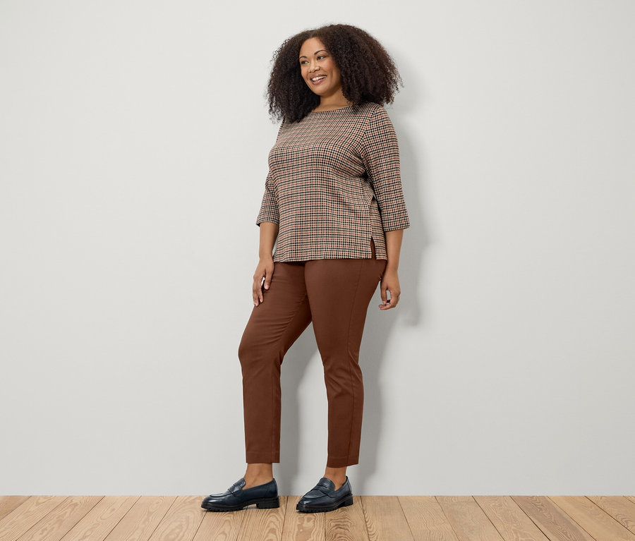 Nő pózol barna 3/4-es ujjú pulóverben, barna nadrágban és fekete cipőben.