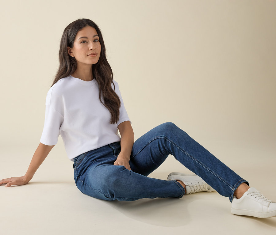 Nő ül a földön Slim-Jeans – Fit »Emma«, középkék farmerben, fehér pólóban és tornacipőben.