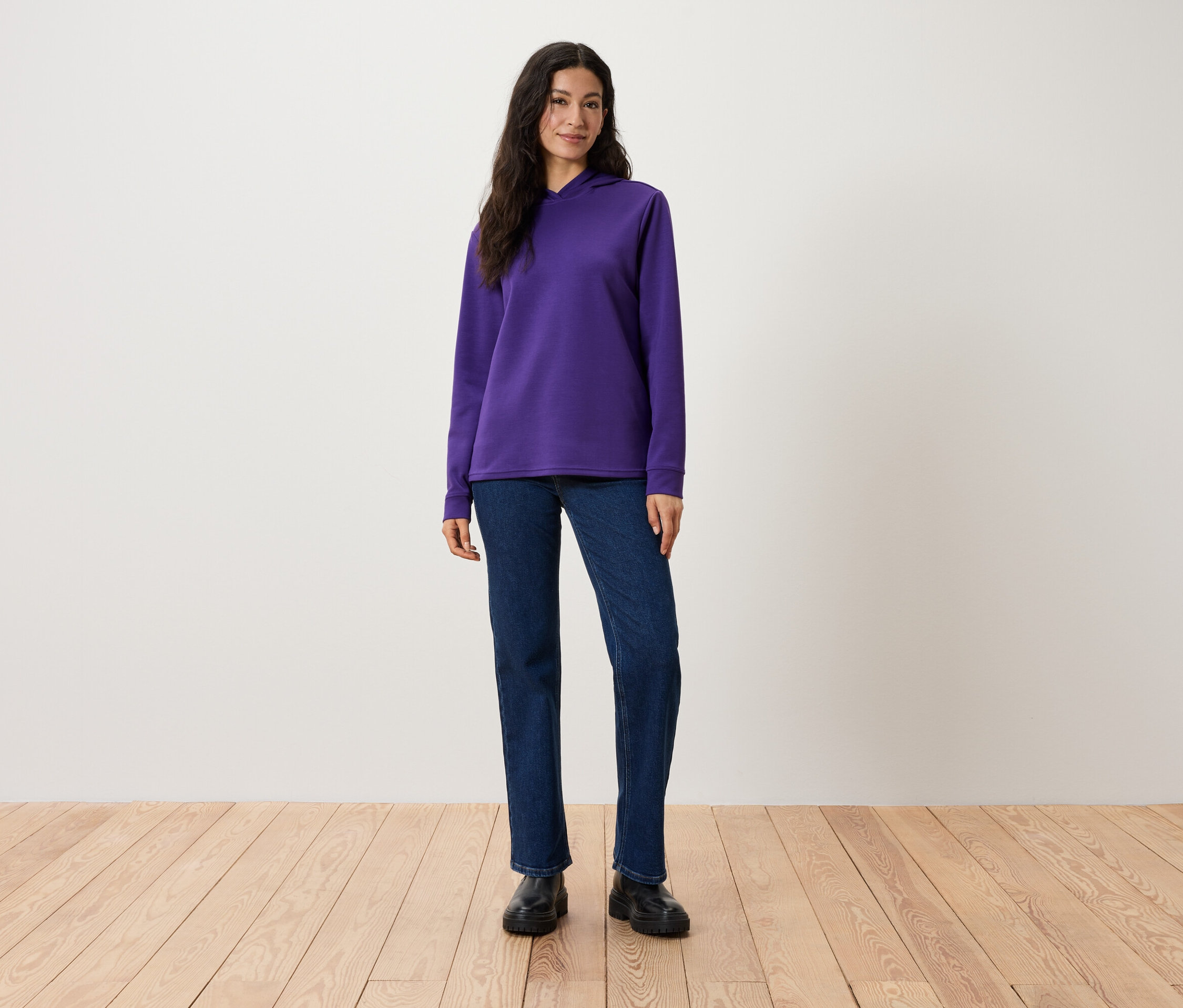 Egy nő áll, lila kapucnis pulóvert, farmert és fekete cipőt visel.