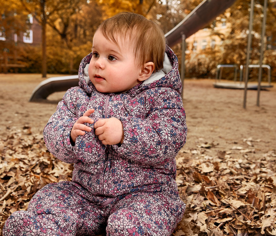 Egy baba ül egy játszótéren a levelek között egy Baby-Winteroverallban.