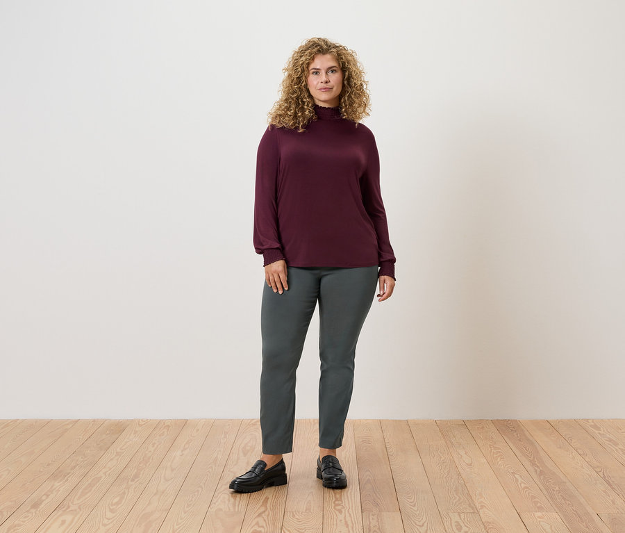 Nő áll a fapadlón, sötétzöld sztreccsnadrágot és bordó hosszú ujjú felsőt visel.