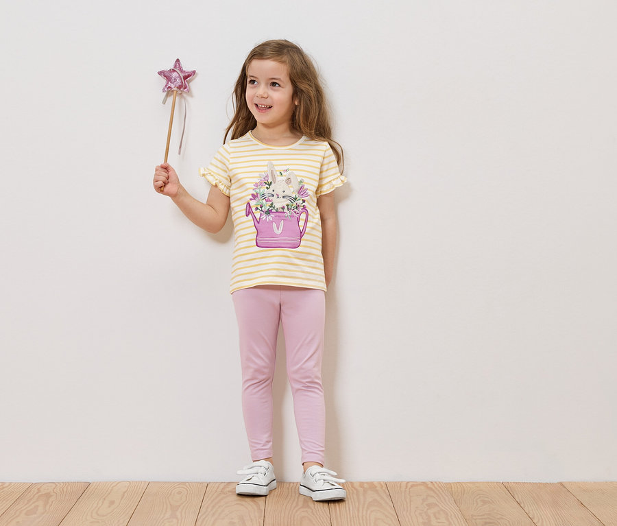 Kislány csillagpálcát tart, csíkos pólót visel, amin virágos locsolókanna van, és rózsaszín leggingset visel.