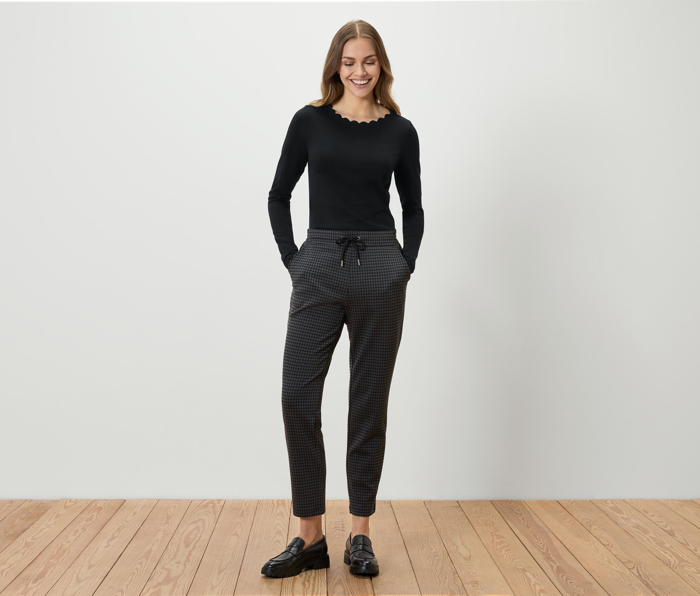 Egy nő áll és mosolyog, fekete hosszú ujjú felsőt, szürke mintás nadrágot és fekete mokaszint visel.