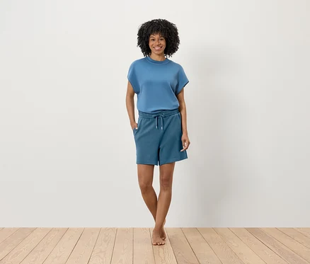 Afro frizurás nő áll a fa padlón egy fehér fal előtt. Kék pólót és rövidnadrágot visel.