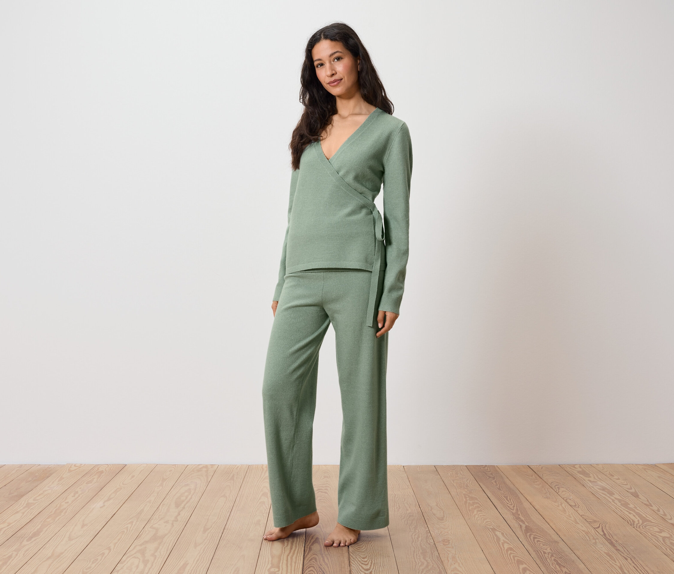 Nő zöld garbóban és nadrágban áll a fapadlón.