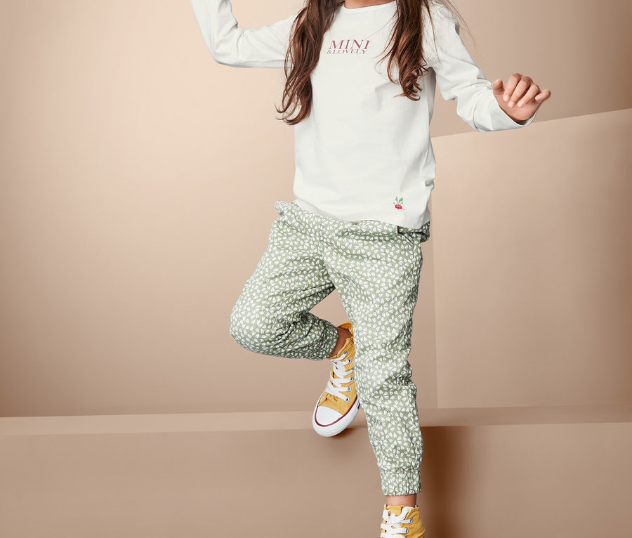 Egy lány pózol fehér, hosszú ujjú felsőben, mintás zöld nadrágban és sárga tornacipőben.